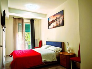 A hotel room with a bed, a nightstand, a red chair, a painting on the wall, and a window with curtains.