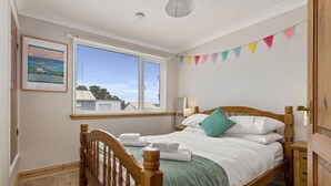 Ein ordentlich bezogenes Bett mit weißen Leinen und einer grünen Tagesdecke, ein hölzernes Kopfteil, ein Nachttisch mit Lampe und ein Fenster mit Jalousien.
