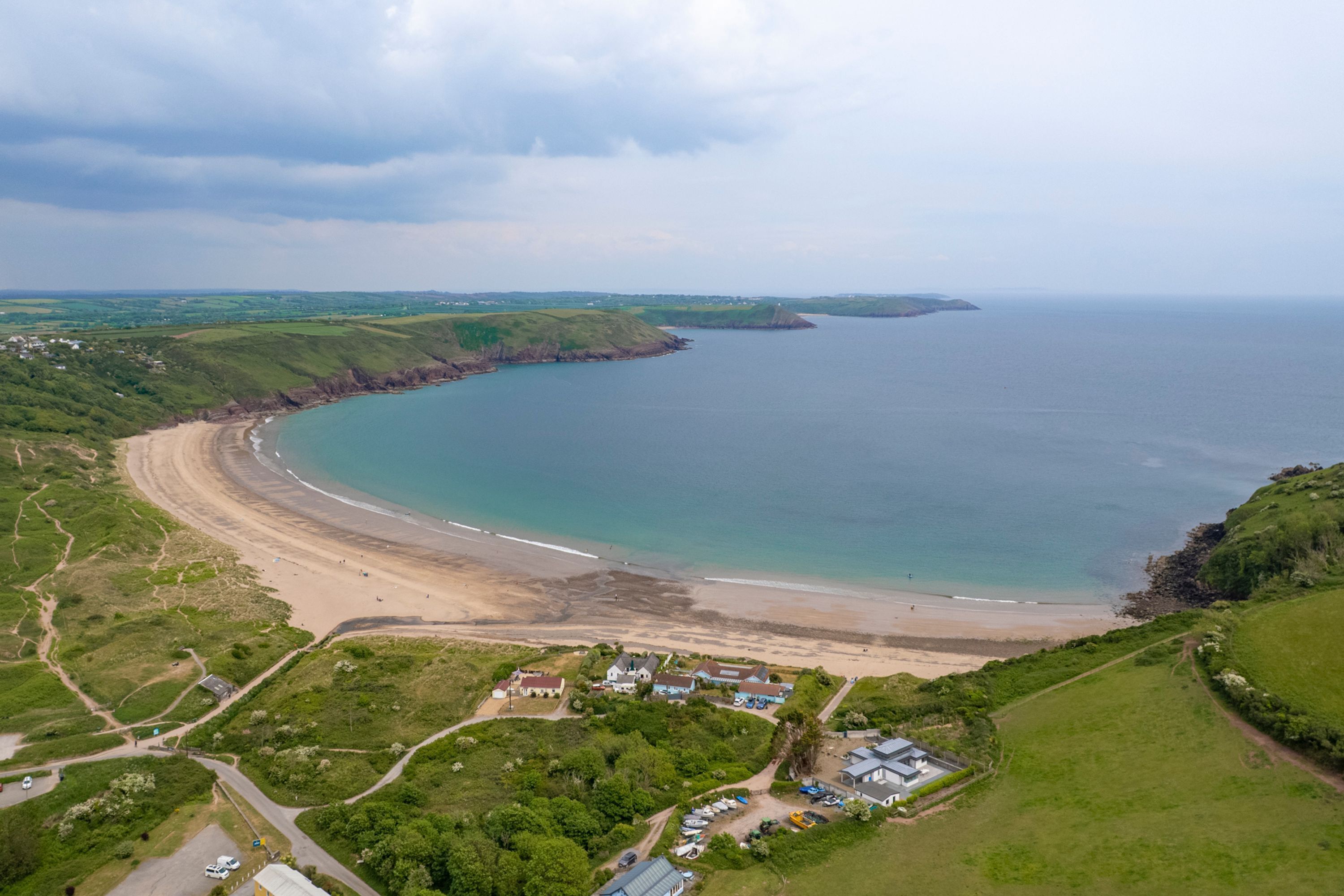 An aerial view of a coastal area with a beach, green hills, and a small cluster of buildings.