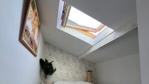 Un dormitorio pequeño y acogedor con techo inclinado y tragaluces, una cama con colcha blanca, un cuadro en la pared y una planta en maceta.