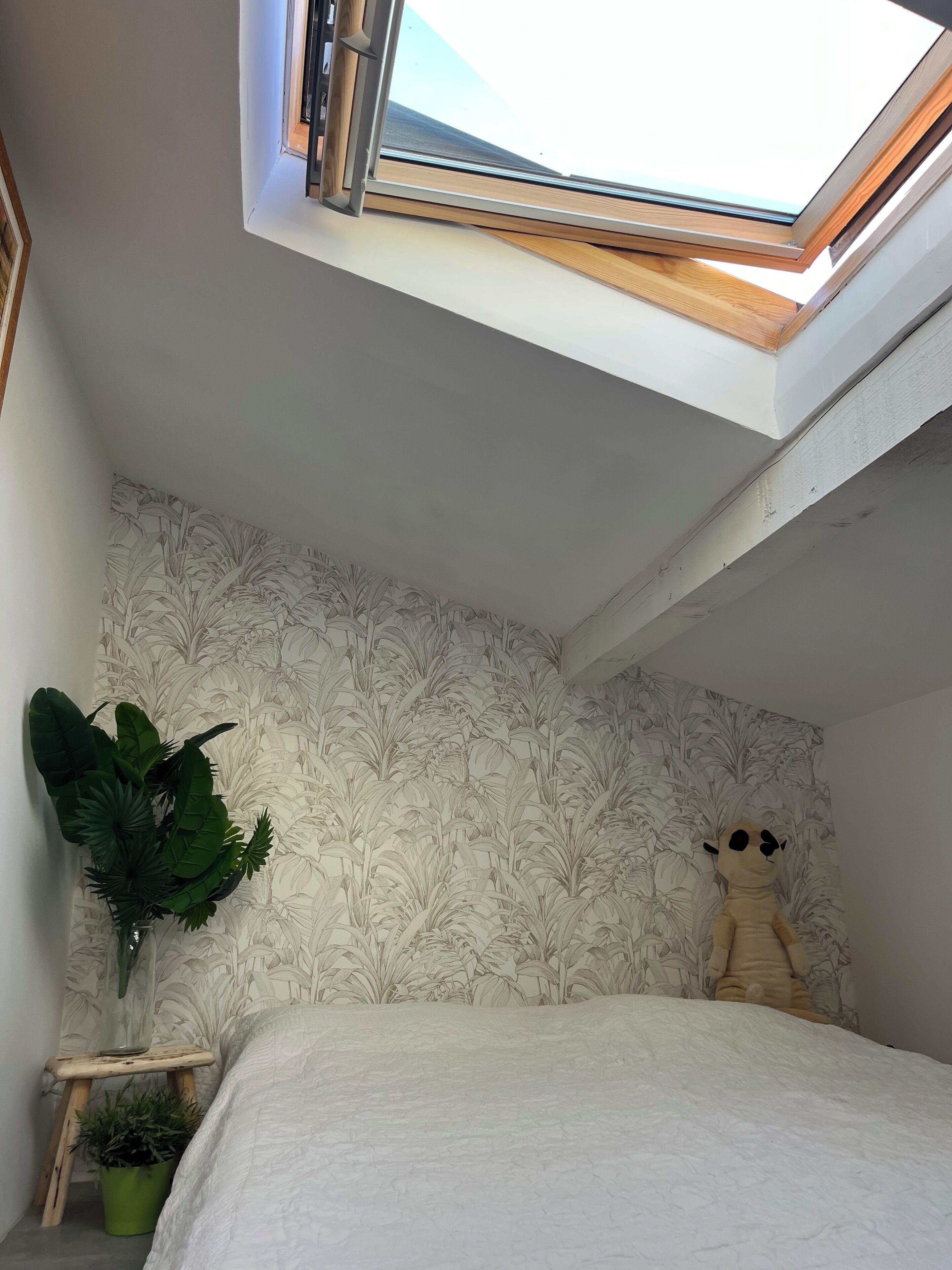 Ein Schlafzimmer mit einem Oberlicht, einem Bett, einer großen Topfpflanze, einem kleinen Hocker und einem Kuscheltier vor einer gemusterten Tapete.