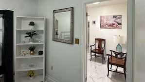 A hallway with a white marble floor, a white shelving unit with decorative items, a mirror, and a wooden chair with a lamp.