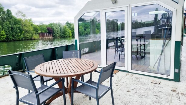 An outdoor deck with a round wooden table and four chairs, overlooking a river with greenery and a dock in the background.