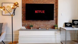 A modern living room with a brick wall, a mounted TV displaying 'NETFLIX,' a floor lamp, a white console table with decorative items, and a desk with a laptop and a router.