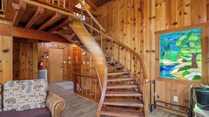 Interior de madera con una escalera espiral, un sofá con cojines de estampado animal y un ventanal con vitrales que representa un paisaje.