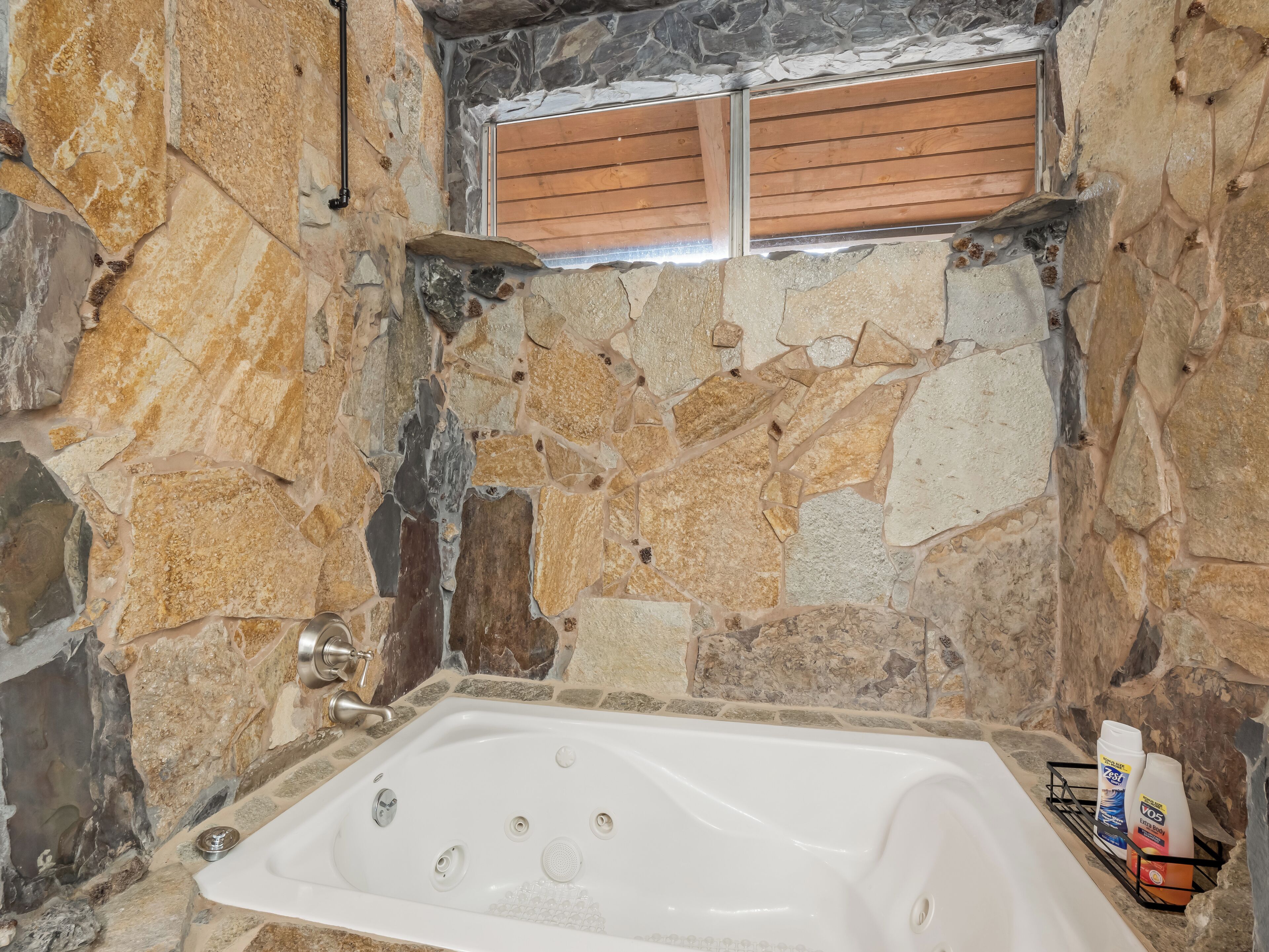 Une salle de bain aux murs de pierre, avec une baignoire, une fenêtre avec des stores en bois et des articles de toilette rangés sur une étagère.