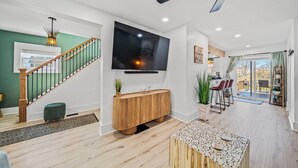 A modern living room with a wooden staircase, a large flat-screen TV, and a dining area with chairs and a table. - Lakeview Modern Stay Near Beaches, GOTL, and Spire (Ashtabula)