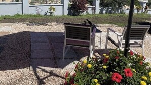 A patio with two wooden chairs, a red umbrella, and a flower bed with red and yellow flowers.