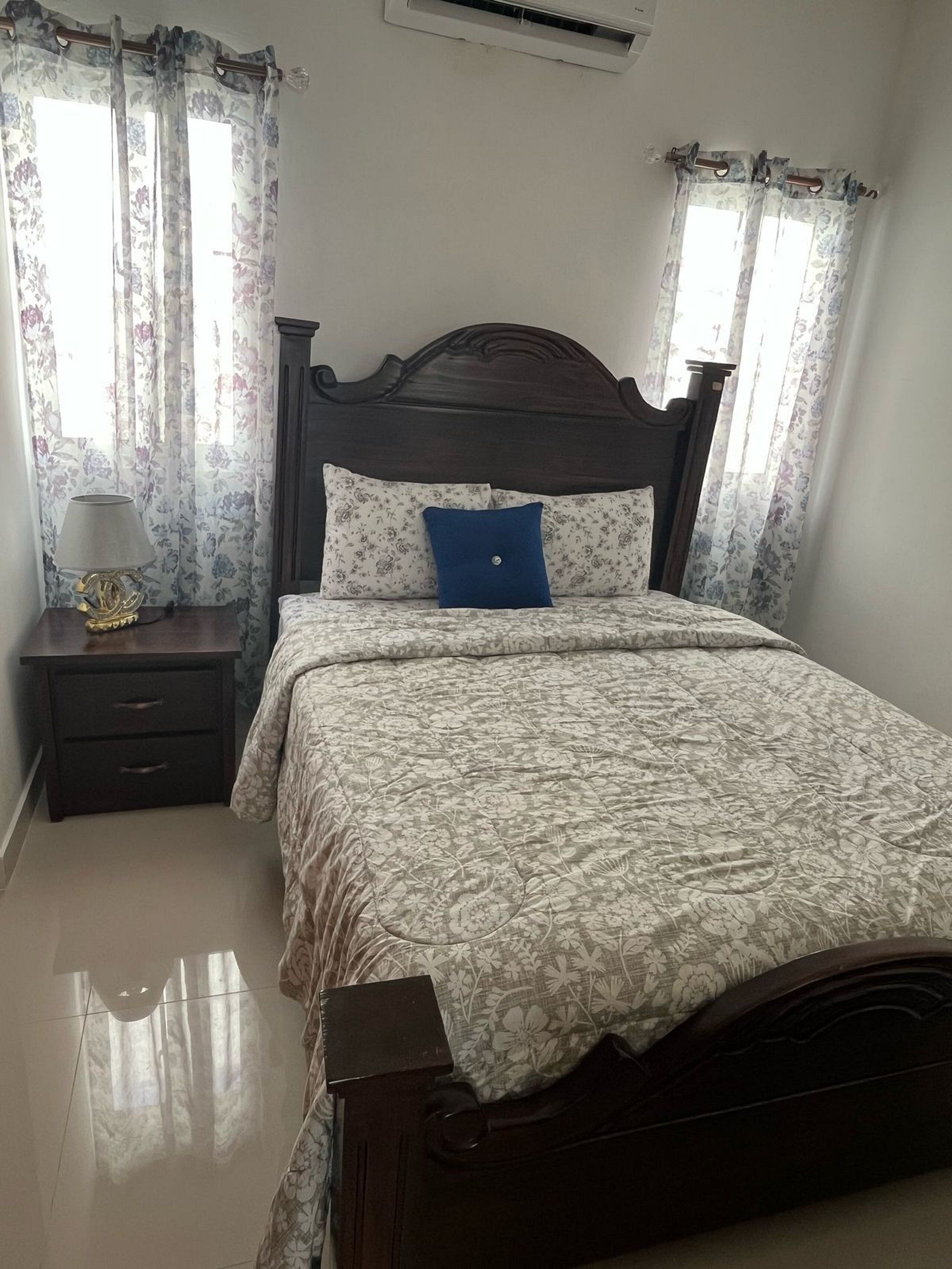 Een slaapkamer met een houten bed, beddengoed met bloemenprint, een nachtkastje met een lamp en gordijnen.