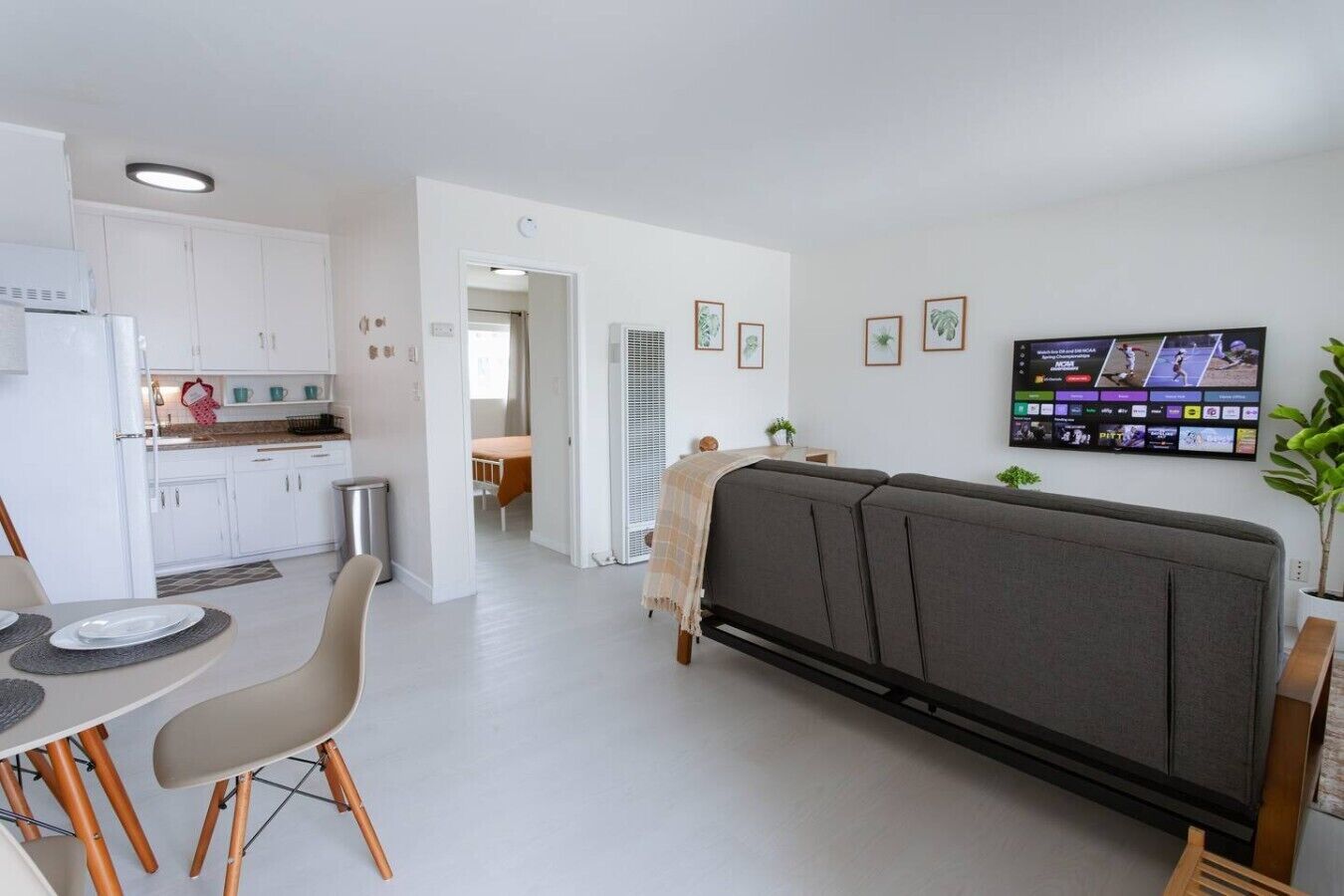 A modern living room with a grey sofa, a dining area with a table and chairs, a kitchen with white cabinets, and a flat-screen TV mounted on the wall.