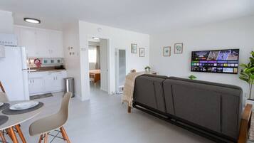 A modern living room with a grey sofa, a dining area with a table and chairs, a kitchen with white cabinets, and a flat-screen TV mounted on the wall.