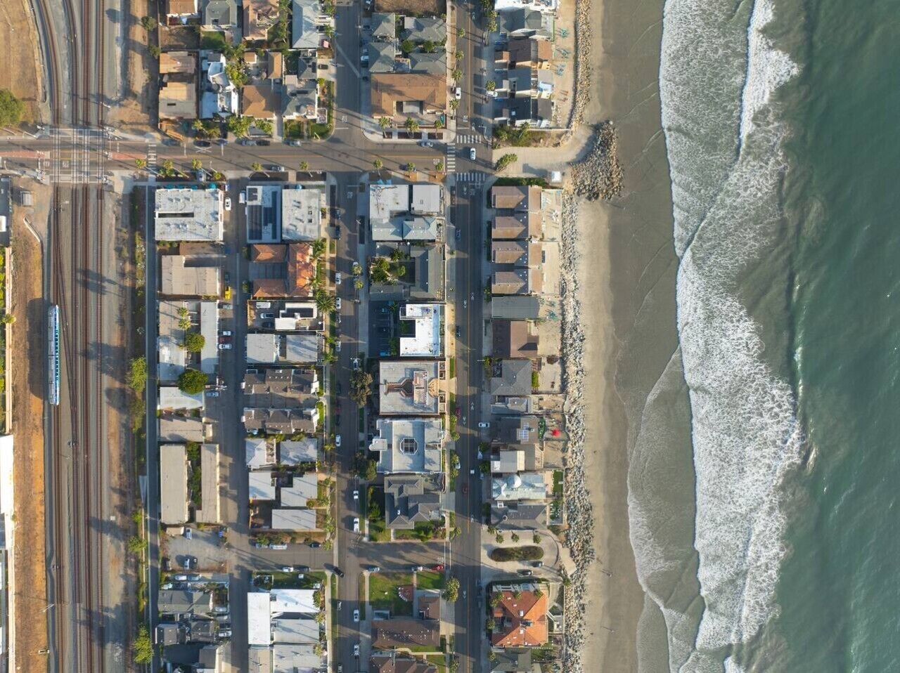 An aerial view of a coastal residential area with houses, a train track, and a beach.