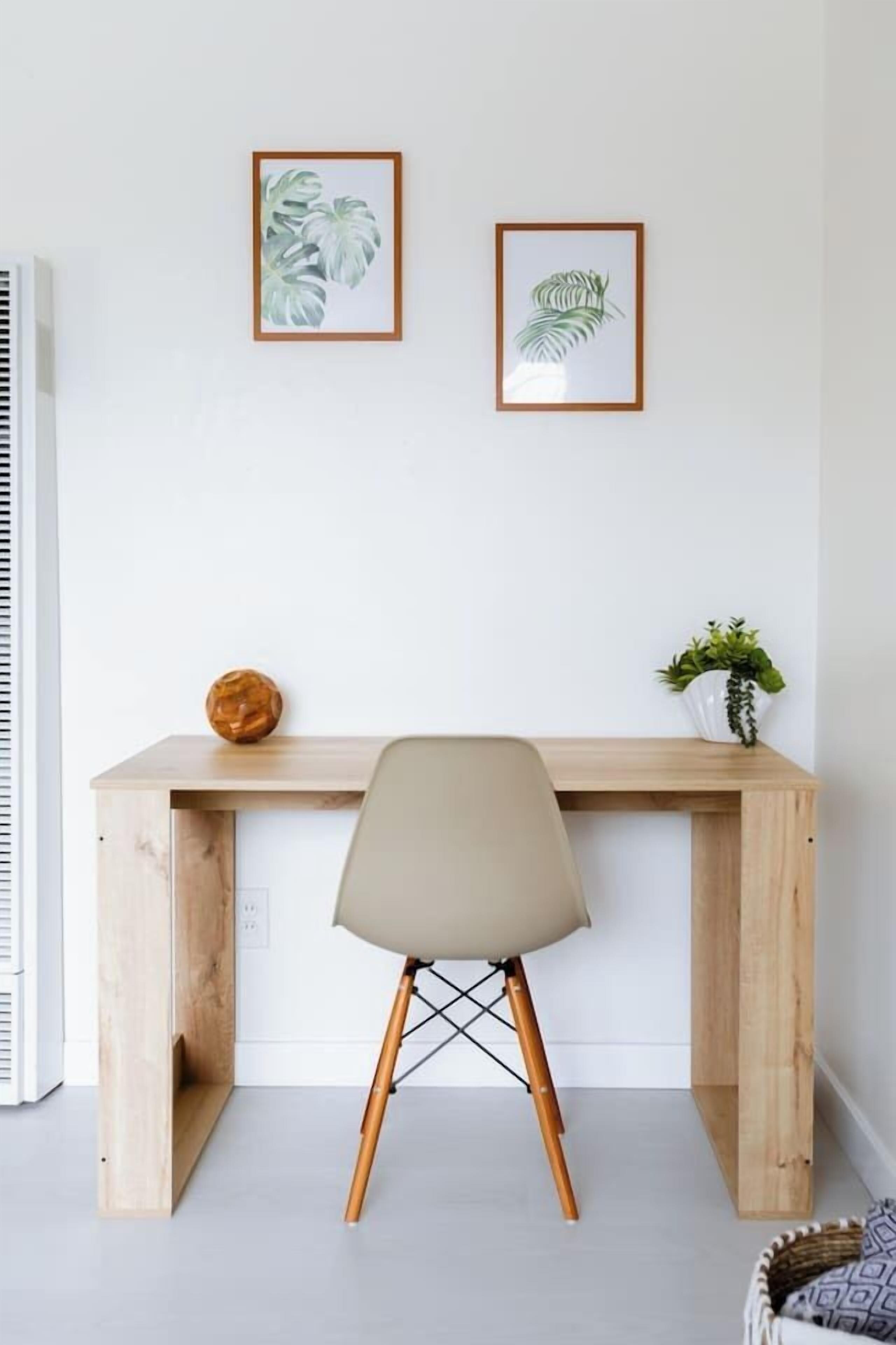 Un escritorio de madera con una silla frente a él, una planta en maceta y dos láminas botánicas enmarcadas en la pared.
