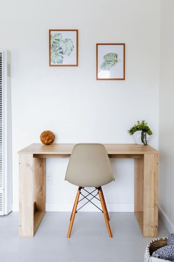 A wooden desk with a chair in front, a potted plant, and two framed botanical prints on the wall.