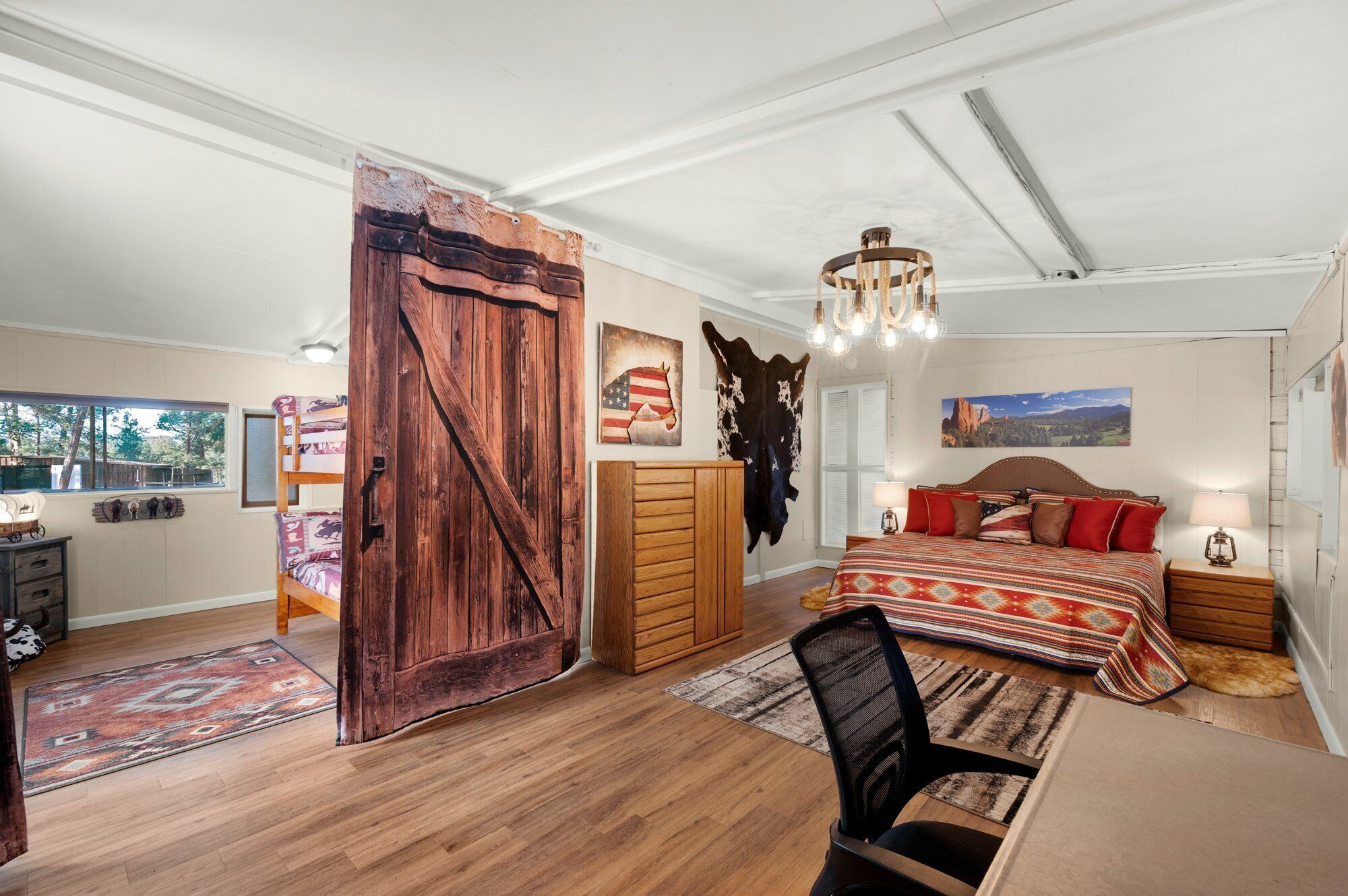 A spacious bedroom with a large wooden door, a bed with patterned bedding, a desk with a chair, and a view of the outdoors.