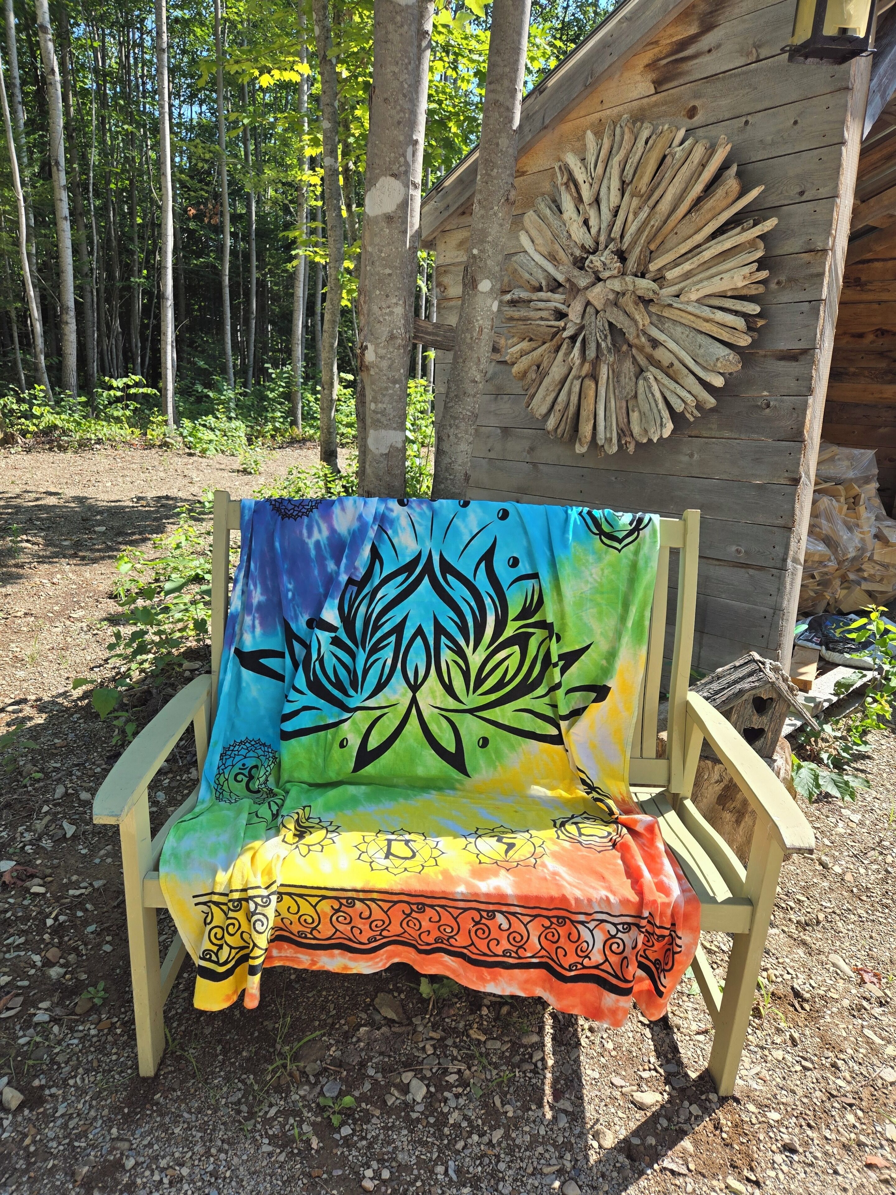 Un banc en bois, recouvert d’une tapisserie aux couleurs vives, se dresse sur fond d’arbres et devant une structure en bois au design original.