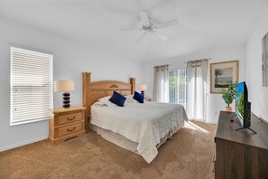 Une chambre à coucher comprenant un lit, des tables de chevet, un ventilateur de plafond, une télévision et une fenêtre avec des stores.