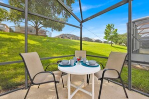 Une terrasse couverte avec une table dressée pour deux personnes, entourée de chaises, donnant sur une pelouse avec des arbres et un bâtiment en arrière-plan.