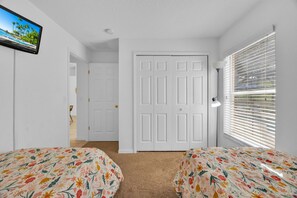 A bedroom with two beds, a TV on the wall, a door, and a window with blinds.