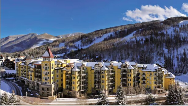Ein mehrgeschossiges Hotel mit gelber Fassade, eingebettet in eine Berglandschaft mit verschneiten Gipfeln.