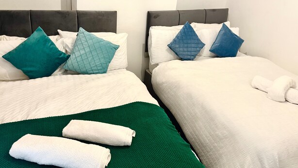 Dos camas de hotel juntas con sábanas blancas, almohadas estampadas en verde azulado y azules, y toallas verdes y blancas.