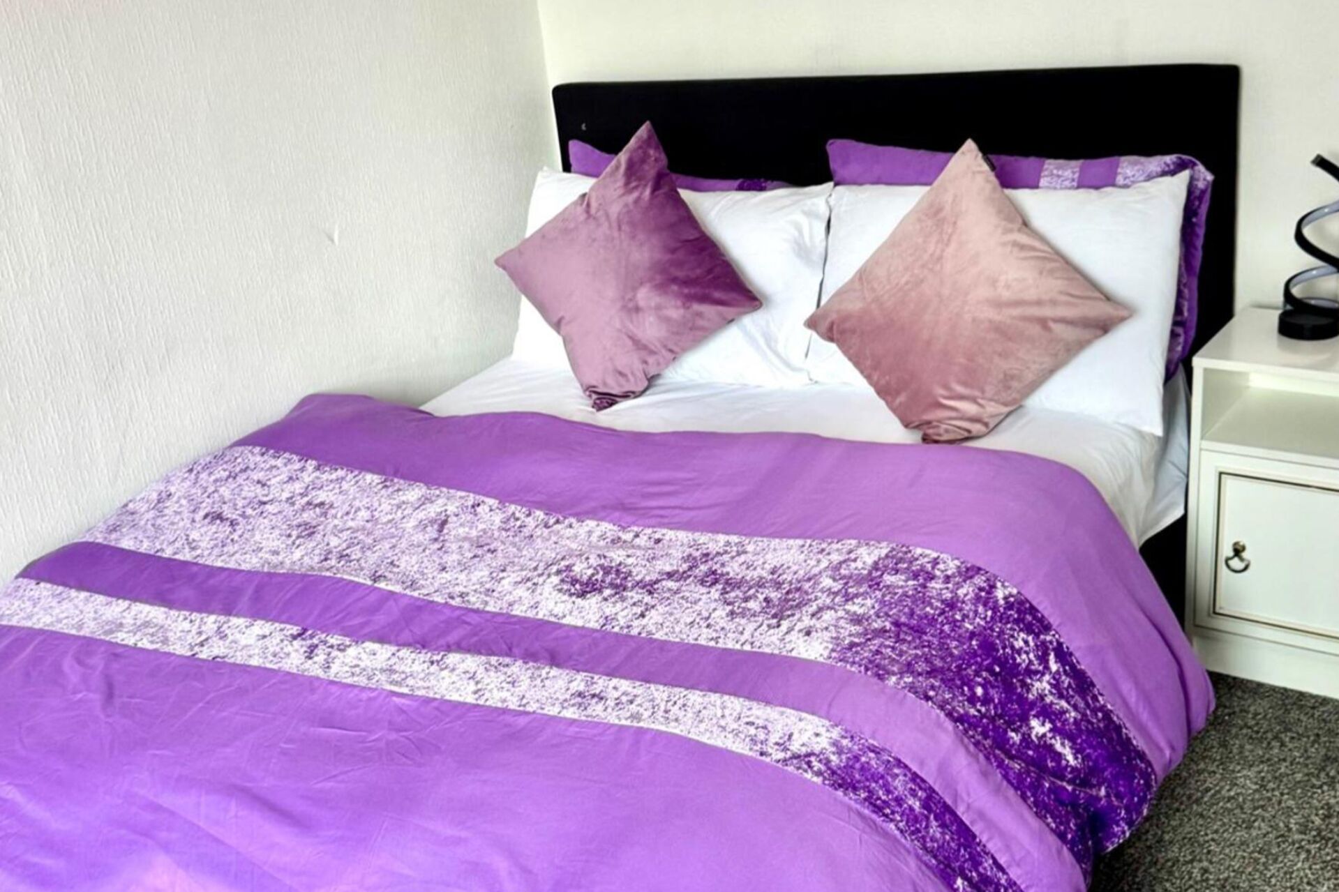 A bed with a purple and white bedspread, two pillows, and a nightstand with a lamp.