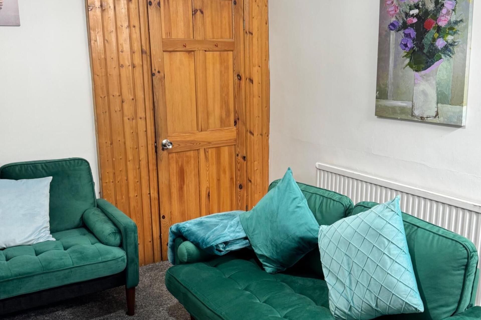 A room with a wooden door, a green sofa with cushions, and a painting of flowers on the wall.