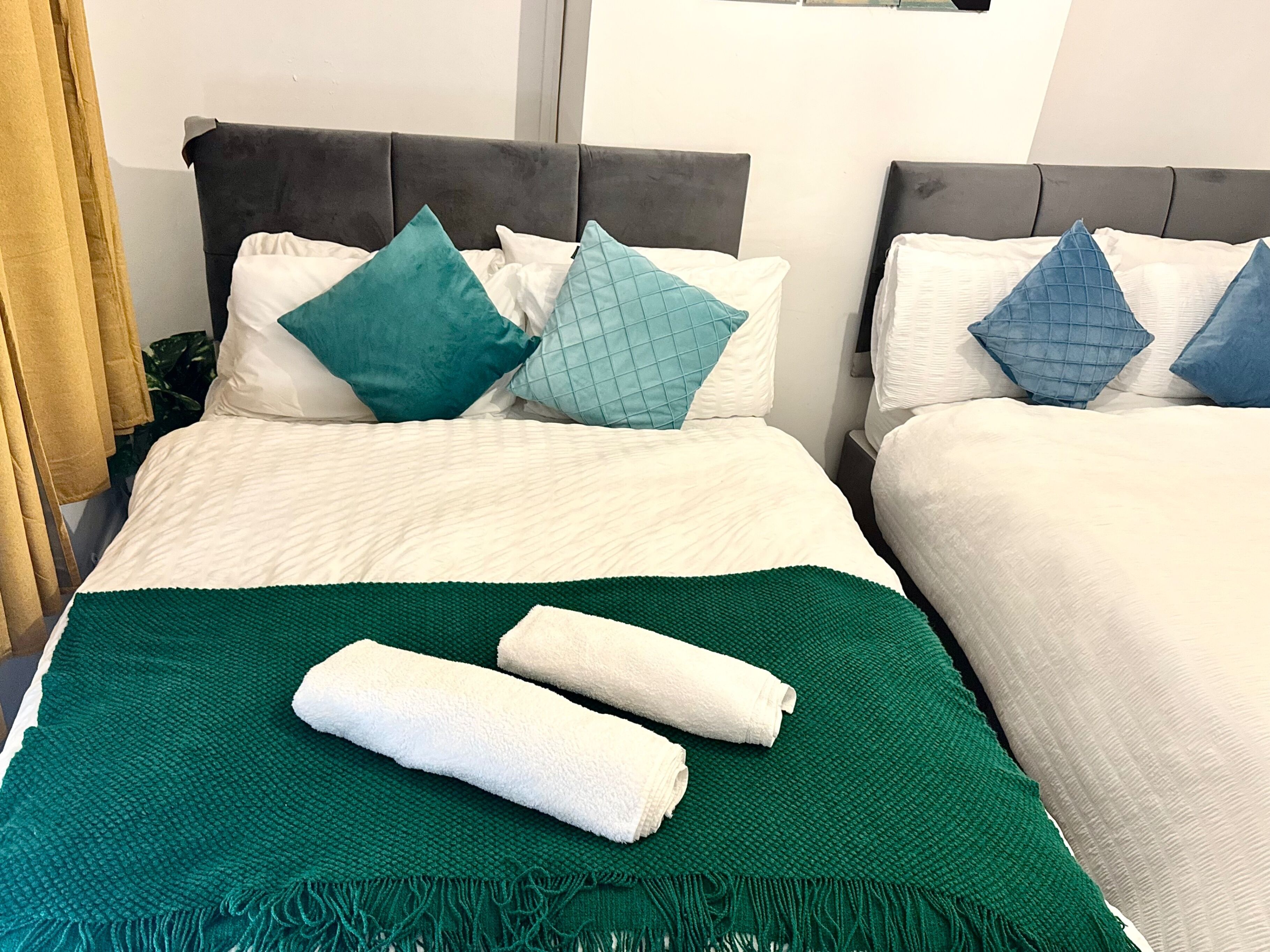 Une chambre d’hôtel avec deux lits, chacun recouvert d’une literie blanche et agrémenté d’oreillers turquoise et bleus. Un plaid vert est posé sur le lit au premier plan.