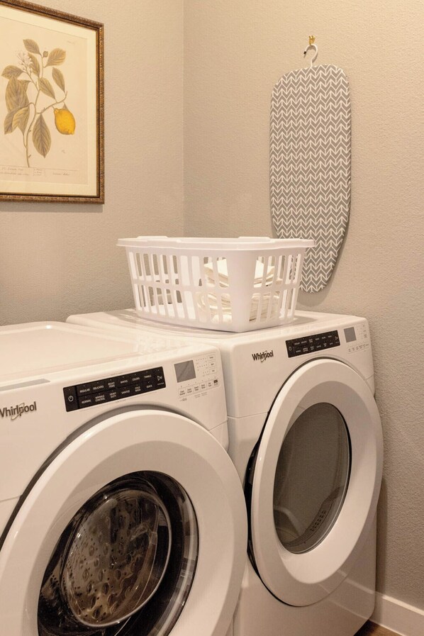 A laundry area with two Whirlpool washing machines, a white laundry basket, and a framed botanical illustration on the wall.