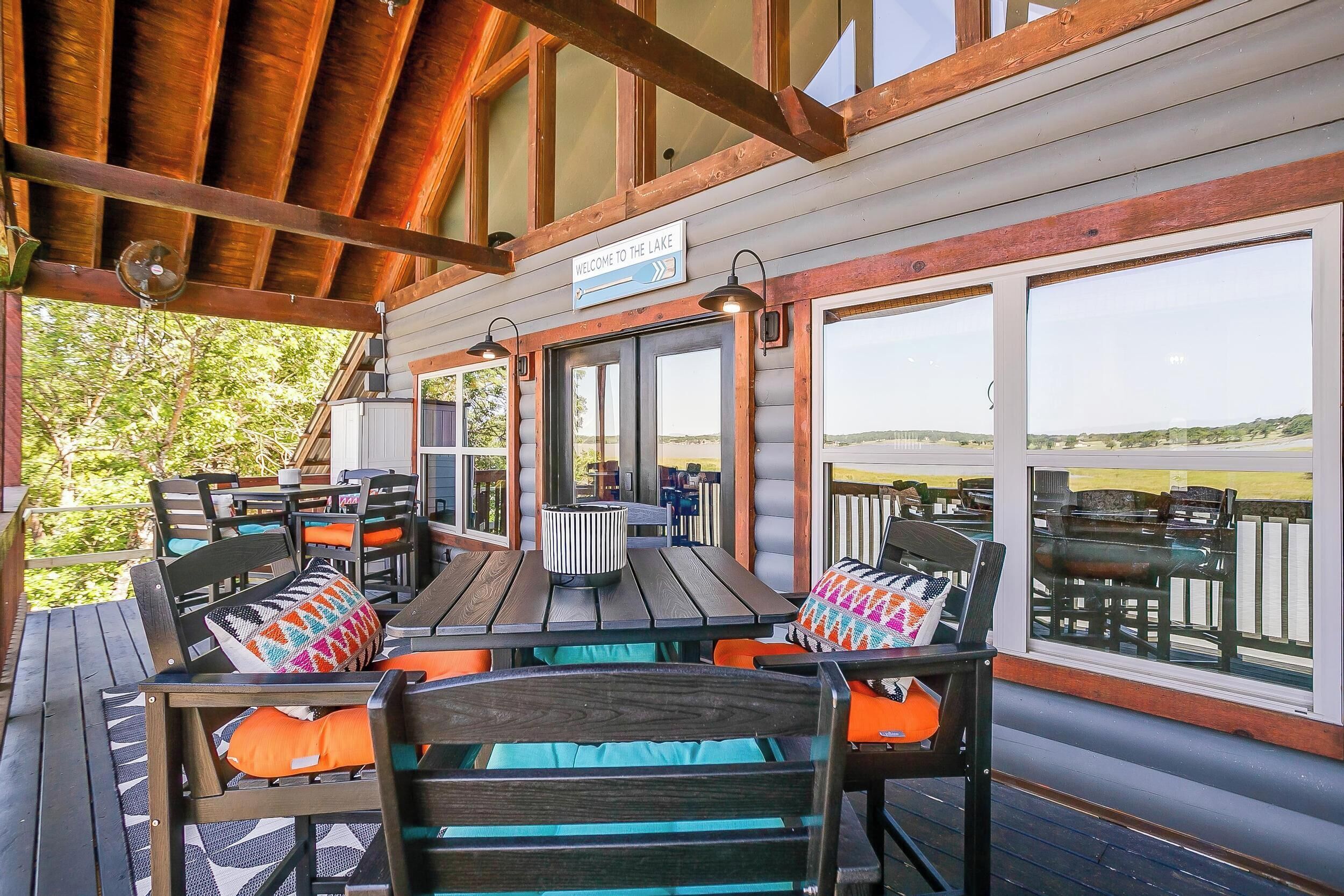 Un espace extérieur aménagé avec des tables et des chaises en bois, protégé par un auvent, une enseigne indiquant « Bienvenue au bord du lac », et de grandes fenêtres offrant une vue sur le lac.