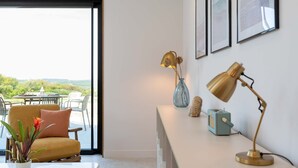 A modern living room with a sofa, a coffee table, a lamp, and a vase with dried flowers. There are framed pictures on the wall and a view of the outdoors through a glass door. - Woodlark -  Luxury Cottages (Bradworthy)