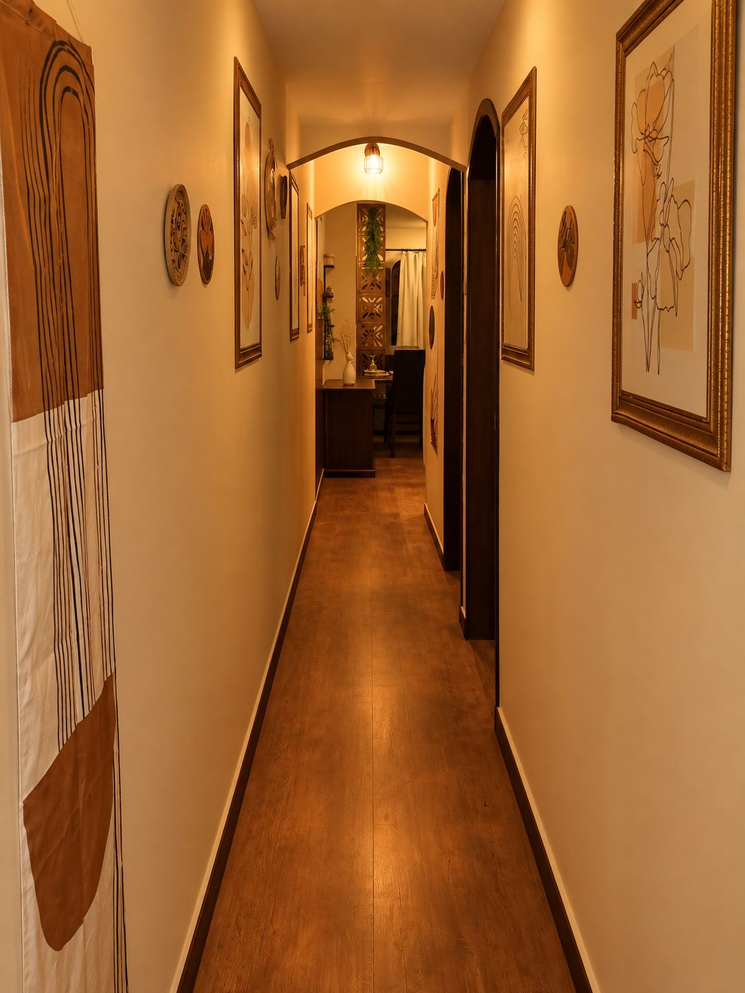 Un pasillo con obras de arte enmarcadas en las paredes, piso de madera y una puerta que da a una habitación con escritorio y silla.