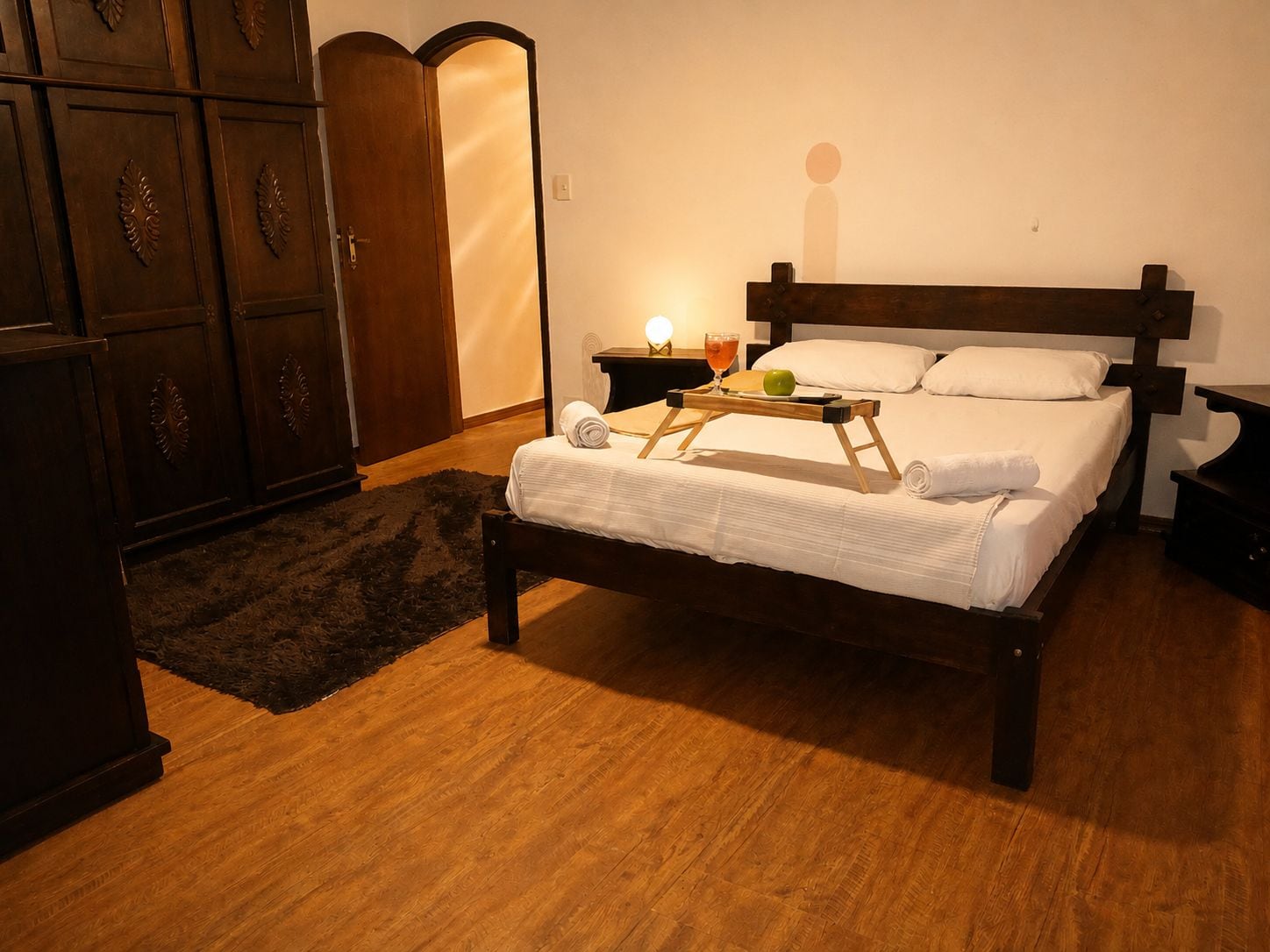 Ein ordentlich bezogenes Bett mit einem hölzernen Kopfteil, ein Nachttisch mit einer Lampe sowie ein Tablett mit einem Getränk und einem Apfel.