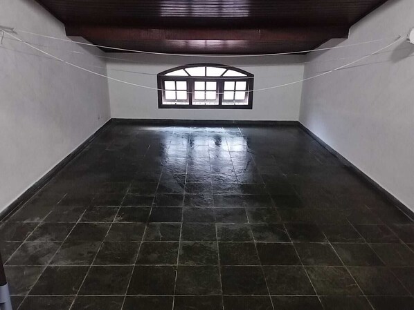 Een kamer met een donkere betegelde vloer, witte muren en een houten plafond. Er is een raam met een zwarte lijst en een witte gordijn.