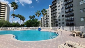 Une piscine avec des chaises longues et une clôture blanche, entourée de palmiers et d’immeubles à plusieurs étages.