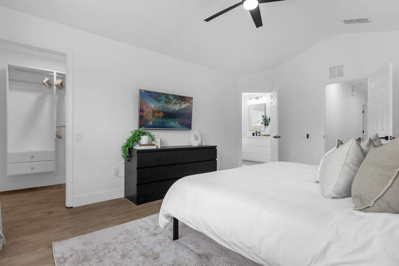 A modern bedroom with a large bed, a dresser, a painting, and a ceiling fan.