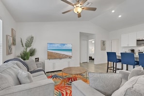 A spacious living room with a ceiling fan, a flat-screen TV, a coffee table, and a kitchen area with blue bar stools.