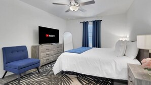 A hotel room with a bed, a TV, a ceiling fan, a blue chair, a wooden dresser, and a patterned rug.