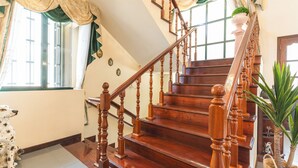 Een houten trap met een balustrade, een hond zittend bij de trap, en een raam met gordijnen.