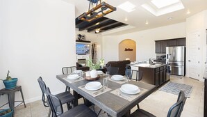 Una cocina moderna con una isla central, un comedor con mesa y sillas de madera, y una sala de estar con sofá y televisor.