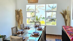 A dining area with a long table set for six, large windows with a view of palm trees, and a woven ceiling light fixture. - Casa de playa a 4 cuadras del mar (Puerto Colombia)