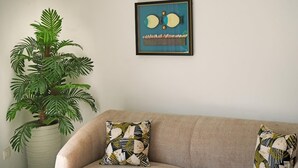 Un sofá color beige con cojines estampados, una obra de arte enmarcada encima y una planta grande en maceta a la izquierda.
