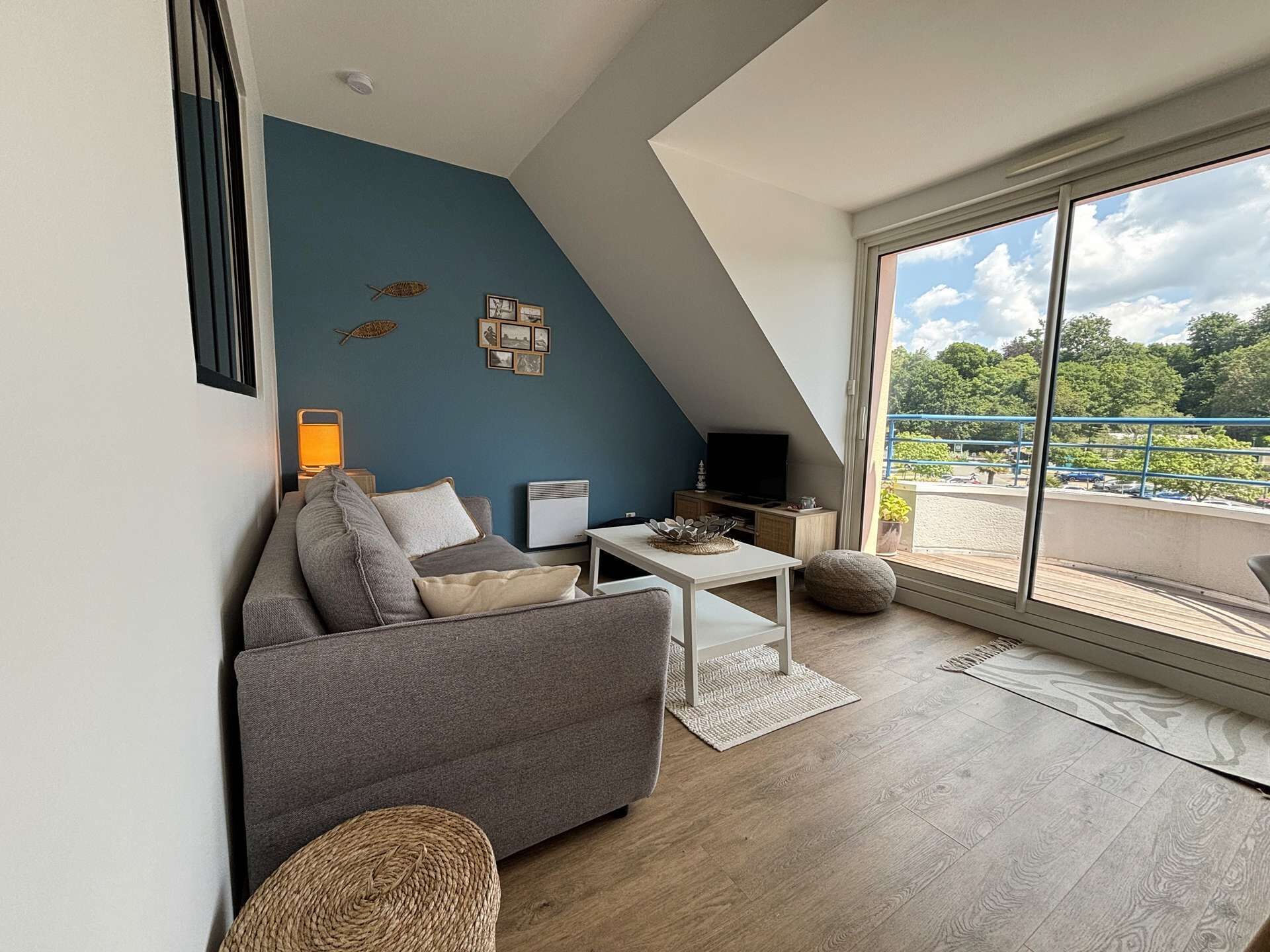 Un salon moderne avec un canapé gris, une table basse blanche et un téléviseur posé sur un meuble en bois. Une grande fenêtre offre une vue sur la verdure et donne accès à un balcon.