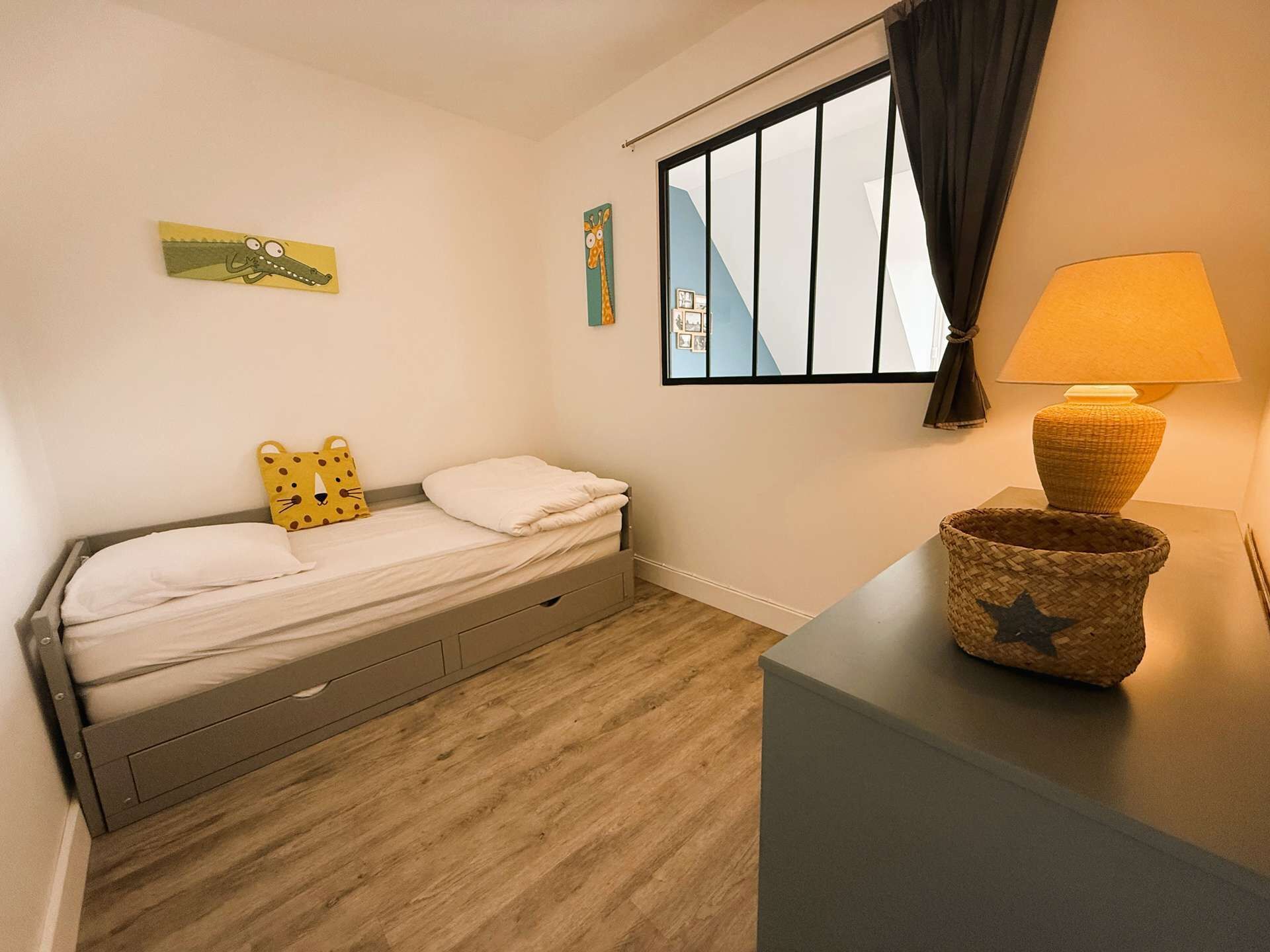 Une petite chambre à coucher, bien rangée, avec un lit simple, une lampe de chevet et une fenêtre avec des rideaux.