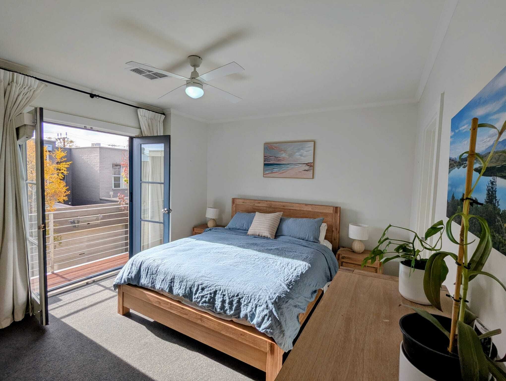 A bedroom with a bed, a ceiling fan, a window with curtains, a painting, and a potted plant.