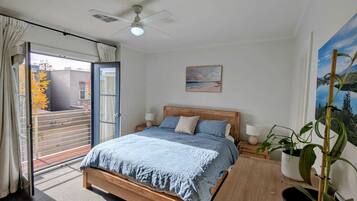 A bedroom with a bed, a ceiling fan, a window with curtains, a painting, and a potted plant.