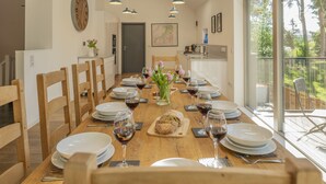 A dining area with a wooden table set for a meal, a clock on the wall, and a view of the outdoors through large windows.