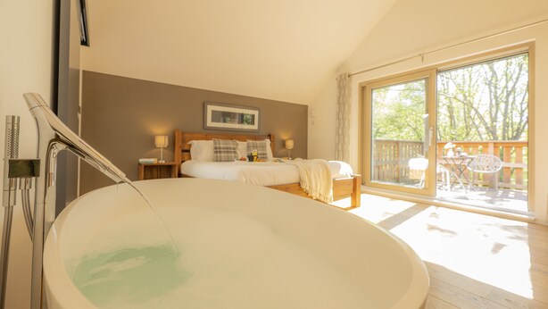 A bedroom with a large bathtub, a bed with pillows, a wooden floor, and a balcony with a view of trees.