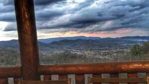 A table with two chairs and two potted plants on a balcony overlooking a cityscape and mountains under a cloudy sky.