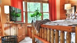 A wooden bedroom with a bed, a TV, a plant, and a window with curtains.